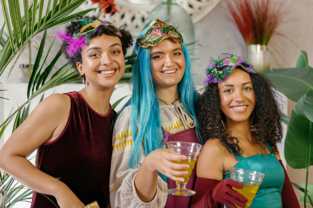 Three women in vibrant masquerade masks and colorful wigs enjoying a fun indoor celebration.